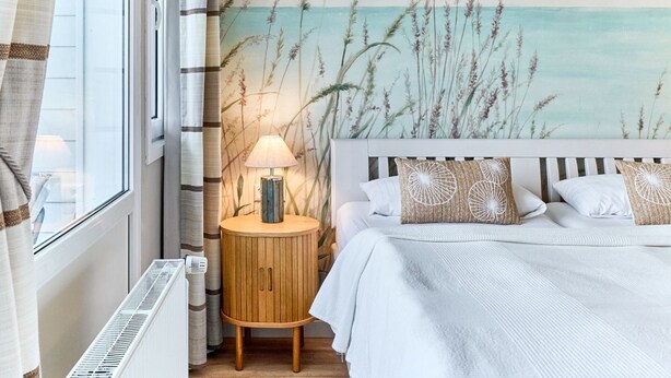 Un dormitorio con cama, lámpara de noche, mesita de noche redonda de madera y un mural en la pared con pastos altos.