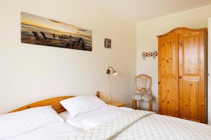 Une chambre à coucher comprenant un lit, une armoire en bois, une chaise, une lampe fixée au mur et un tableau représentant un coucher de soleil sur l’océan.