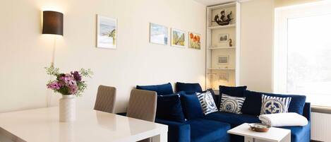 Un salón moderno con una mesa de comedor blanca, un sofá azul y una pequeña mesita auxiliar blanca. Hay obras de arte enmarcadas en la pared y un jarrón con flores sobre la mesa.