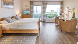 Un dormitorio con cama, mesitas de noche, televisor, ventana con cortinas, zona de comedor con mesa y sillas, y vistas al exterior con vegetación.