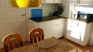 Een compacte keuken met witte kasten, een blauw aanrecht en een magnetron. Er staat een houten tafel met stoelen en geweven placemats.