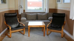 Een woonkamer met een bank, twee leren fauteuils, een houten tafel en een raam met gordijnen.