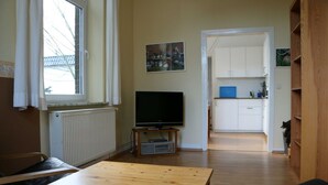 Een kamer met een houten tafel, een televisie op een standaard, een radiator, een raam met gordijnen, en een keukenhoek met kastjes en een blauw aanrecht.