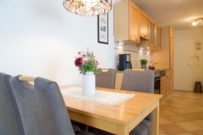 A dining area with a wooden table, grey upholstered chairs, a vase with flowers, and a kitchen in the background with wooden cabinets and a coffee machine.