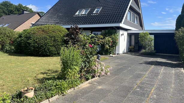 Ett modernt hus i två våningar med ett sluttande tak, ett garage och en välskött trädgård med olika växter och blommor.