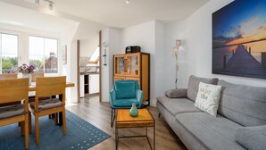 A modern living room with a grey sofa, teal armchair, wooden dining table, and a large painting of a sunset. - (TRA7) Holiday apartment "Reichelt" - Holiday apartment "Reichelt" up to 4 persons (Timmendorfer Strand)