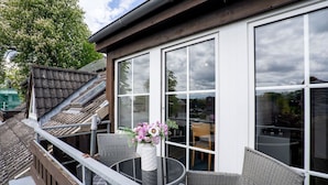 A balcony with wicker furniture, a table with a vase of flowers, and a large window overlooking a residential area. - (TRA7) Holiday apartment "Reichelt" - Holiday apartment "Reichelt" up to 4 persons (Timmendorfer Strand)
