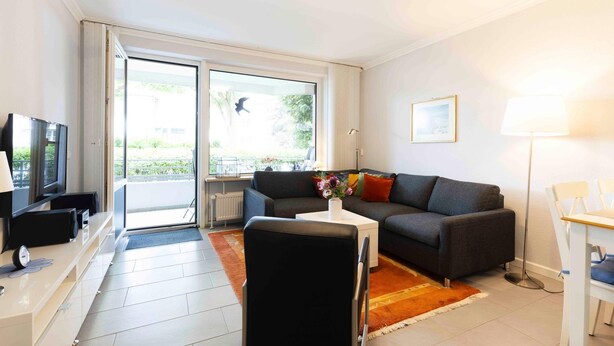 A modern living room with a sofa, a coffee table, a TV, and a dining area with chairs. - (TRA4) Holiday apartment "Hubertus "F*** - Holiday apartment "Hubertus" up to 3 persons (Timmendorfer Strand)