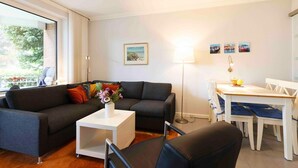 A modern living room with a dark grey sofa, a white coffee table, and a dining table with white chairs. There is a floor lamp, a framed picture on the wall, and a window with curtains. - (TRA4) Holiday apartment "Hubertus "F*** - Holiday apartment "Hubertus" up to 3 persons (Timmendorfer Strand)