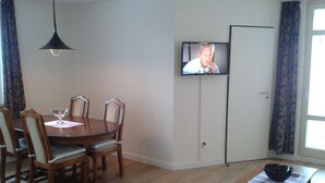 A dining area with a wooden table and chairs, a television mounted on the wall, and a window with curtains. - (STR123b) Holiday apartment "Strandhotel" - App. 24 - Holiday apartment "Strandhotel" - (Timmendorfer Strand)