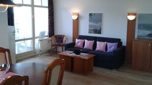 A living room with a sofa, a dining table, and a chair. There is a window with curtains, a radiator, and a painting on the wall. - (STR123b) Holiday apartment "Strandhotel" - App. 24 - Holiday apartment "Strandhotel" - (Timmendorfer Strand)