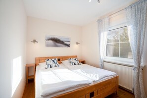 A bedroom with a wooden bed, two bedside tables, a nightstand, a window with curtains, and a wall-mounted picture.