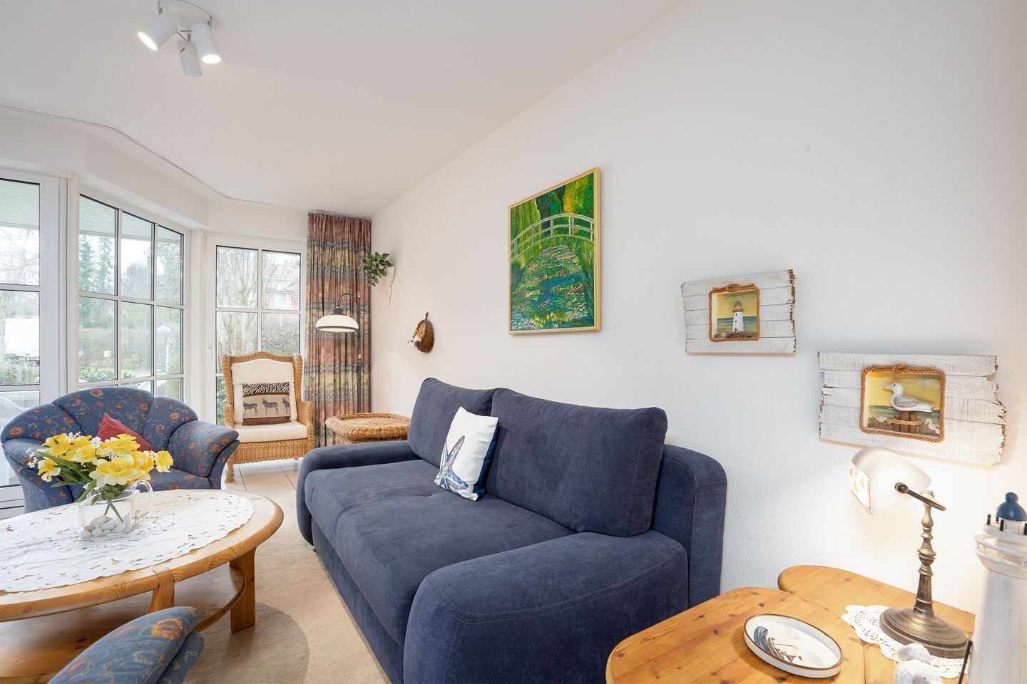 A living room with a blue sofa, a round wooden coffee table, a blue armchair, and a wicker chair. There is a painting on the wall, a lamp, and a framed picture.