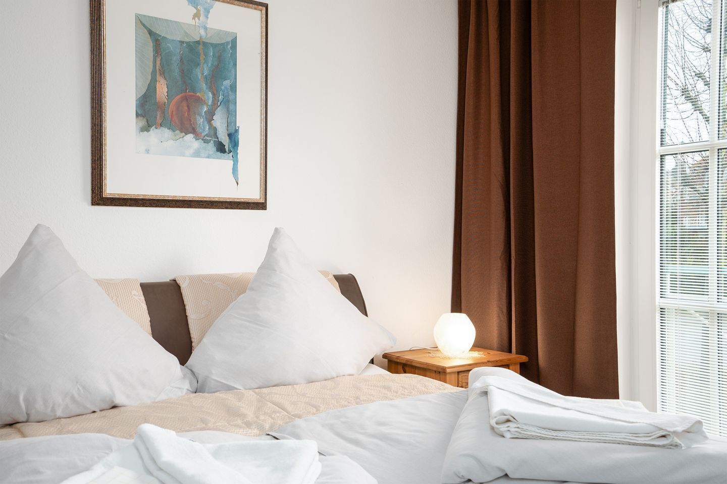 A neatly made bed with white linens, a bedside lamp, and a framed abstract painting on the wall.