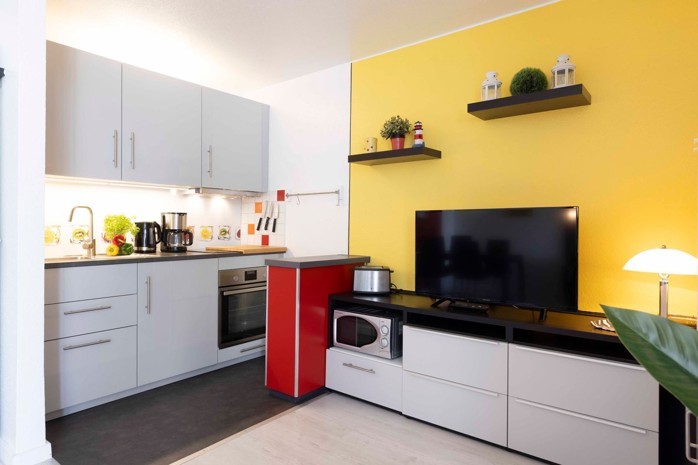 Une cuisine compacte avec des meubles blancs, un mur d’accent rouge, un téléviseur et une petite étagère ornée d’objets décoratifs.