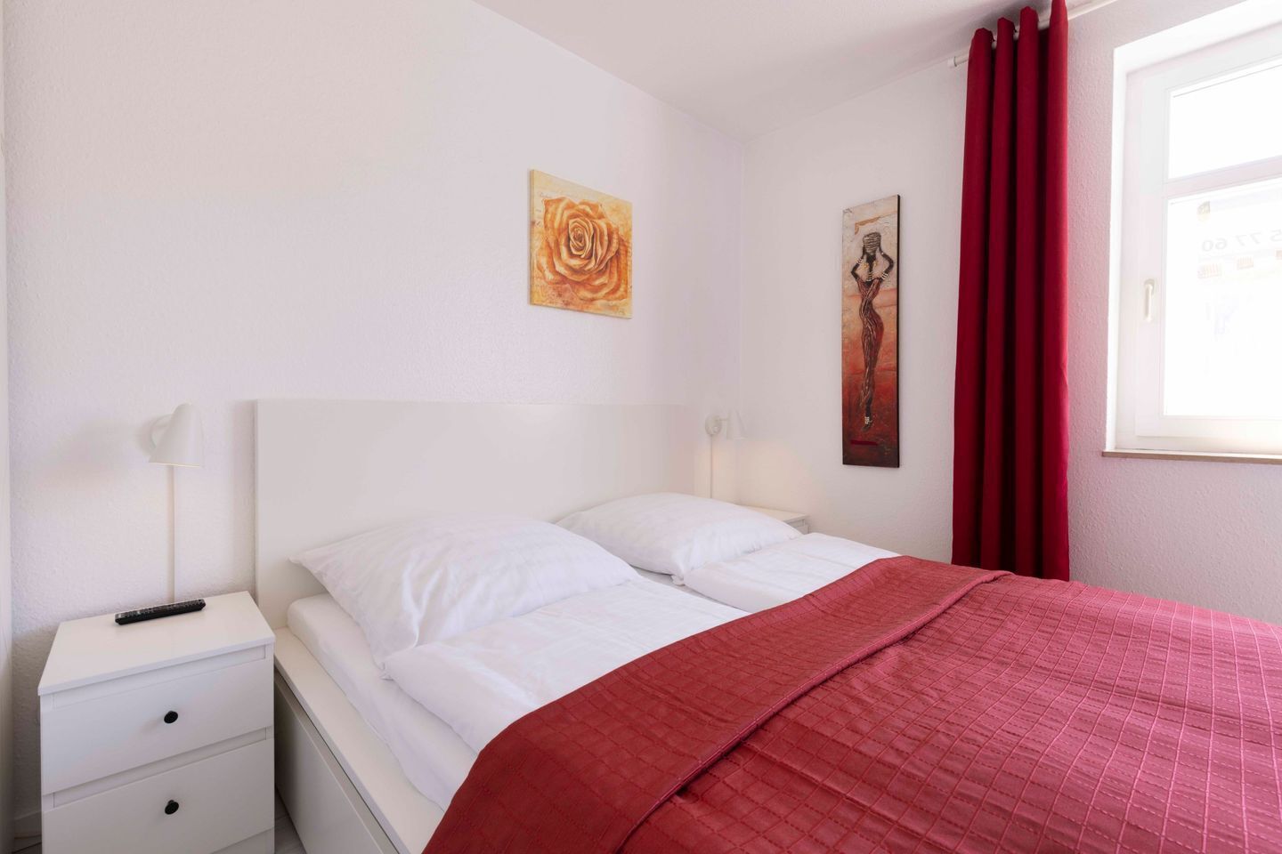 A bedroom with a bed, white nightstands, a red blanket, and two wall art pieces.