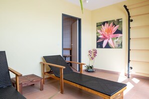 Una habitación con una silla de descanso de madera, una mesita pequeña, una orquídea en maceta y un tendedero de pared.