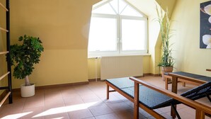 Une pièce baignée de lumière, avec un plafond en pente, une grande fenêtre, un radiateur, une plante en pot et un banc en bois.
