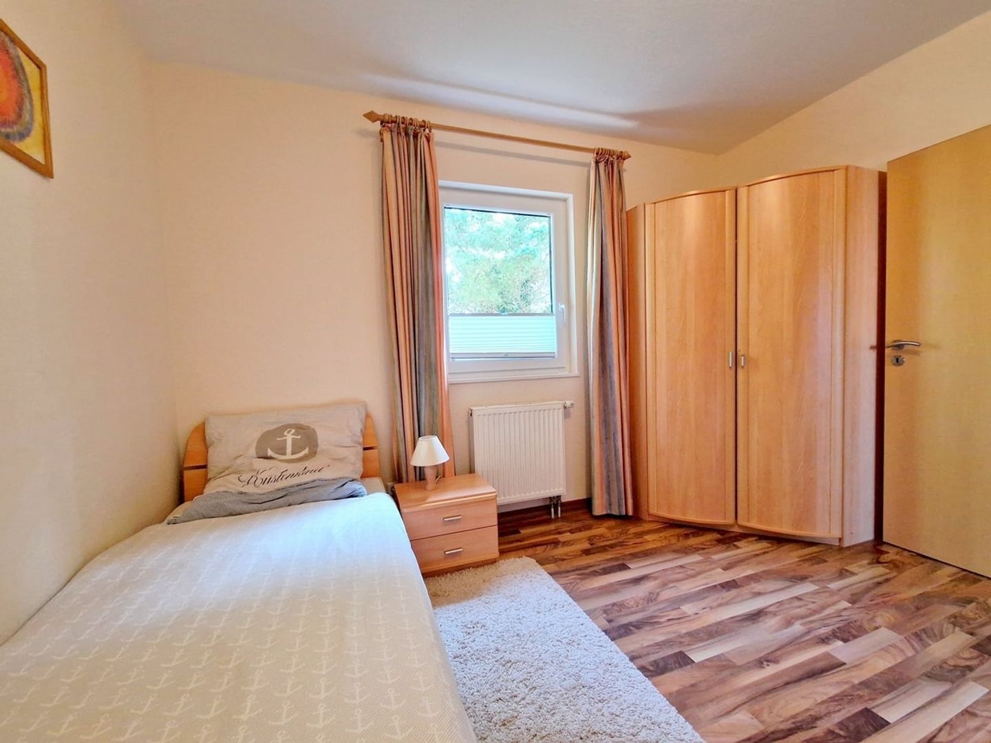 Ein Schlafzimmer mit Bett, Nachttisch, Lampe, Fenster mit Vorhängen, Heizkörper und Kleiderschrank.