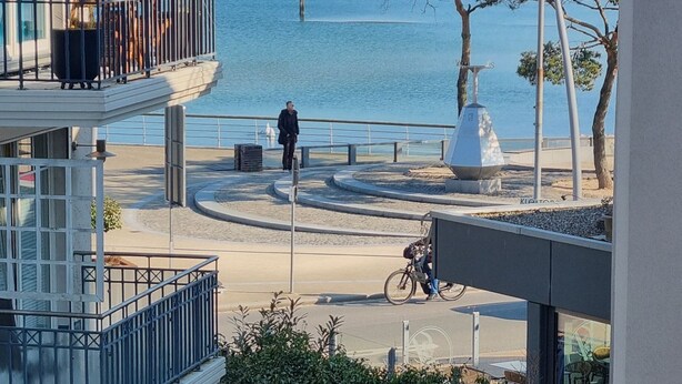 Vista costera desde un balcón con arena dorada y un mar azul cristalino, un árbol solitario y un ciclista.