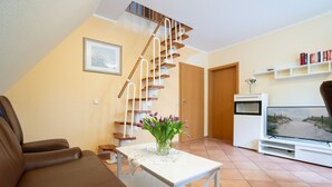 A living room with a brown leather sofa, a white coffee table with a vase of purple flowers, a wooden staircase, a TV on a white stand, and a bookshelf with books. - (STR47a) Holiday apartment "Heitmann" - Holiday apartment "Heitmann" up to 4 persons (Timmendorfer Strand)