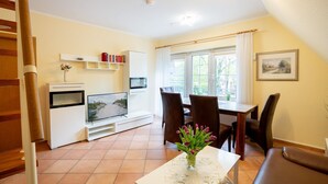 A living room with a TV, a dining table, chairs, a bookshelf, and a window with curtains. - (STR47a) Holiday apartment "Heitmann" - Holiday apartment "Heitmann" up to 4 persons (Timmendorfer Strand)