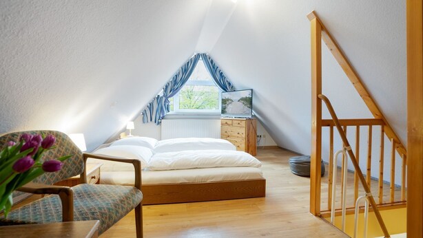 A cozy attic bedroom with a bed, a chair, a desk, and a window with curtains. - (STR47a) Holiday apartment "Heitmann" - Holiday apartment "Heitmann" up to 4 persons (Timmendorfer Strand)