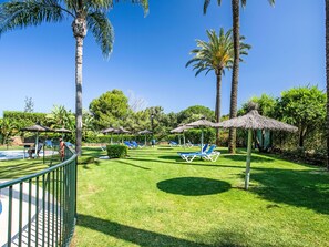 Un espace aménagé au bord de la piscine, avec des chaises longues, des palmiers et une structure dotée d’un toit de chaume.