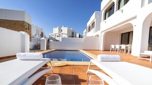 Un área de piscina en la azotea con sillones blancos, una mesa de comedor con vasos, y una vista despejada de los edificios circundantes.