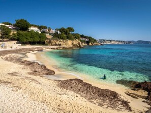 Ein Strand mit klarem Wasser, eine Person beim Schwimmen und Häuser an einem Hügelhang.