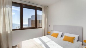 Un dormitorio con una cama grande, dos almohadas, una mesita de noche, una ventana con cortinas y vistas a edificios y al agua.