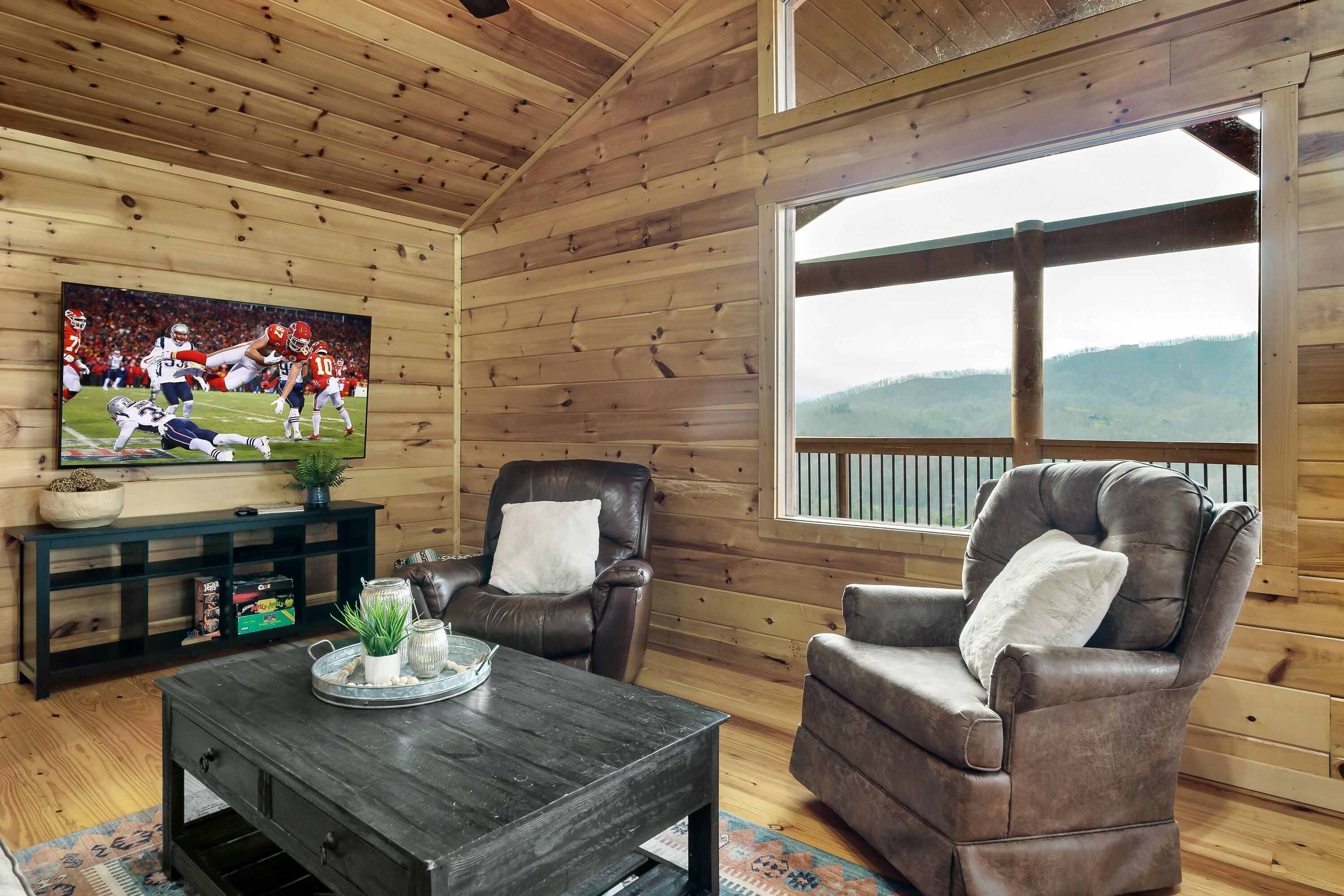 A cozy cabin interior with a wooden ceiling, a large TV displaying a football game, leather chairs, and a view of the outdoors through a large window.