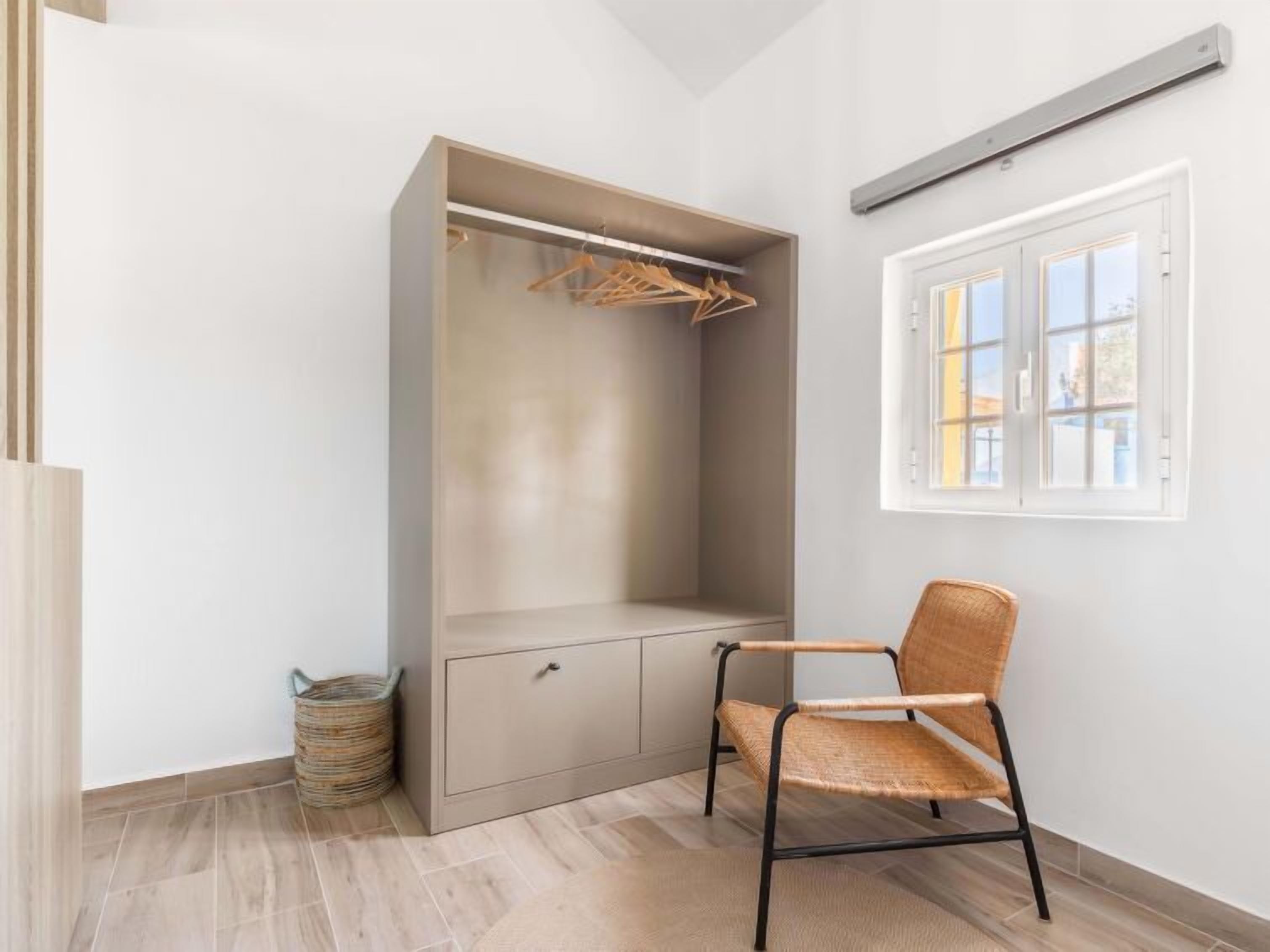 Una habitación con armario empotrado, una silla de mimbre, un cubo de basura pequeño y una ventana con vistas a edificios.