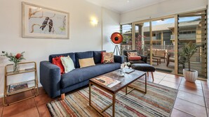 A modern living room with a blue sofa, a coffee table, a framed artwork, and a large window. - The Parap Markets Stay | Walk to Markets & Cafés (Parap)