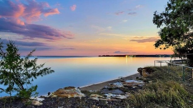 A calm lake at sunset with a rocky shoreline and a distant shoreline with lights. - The Parap Markets Stay | Walk to Markets & Cafés (Parap)