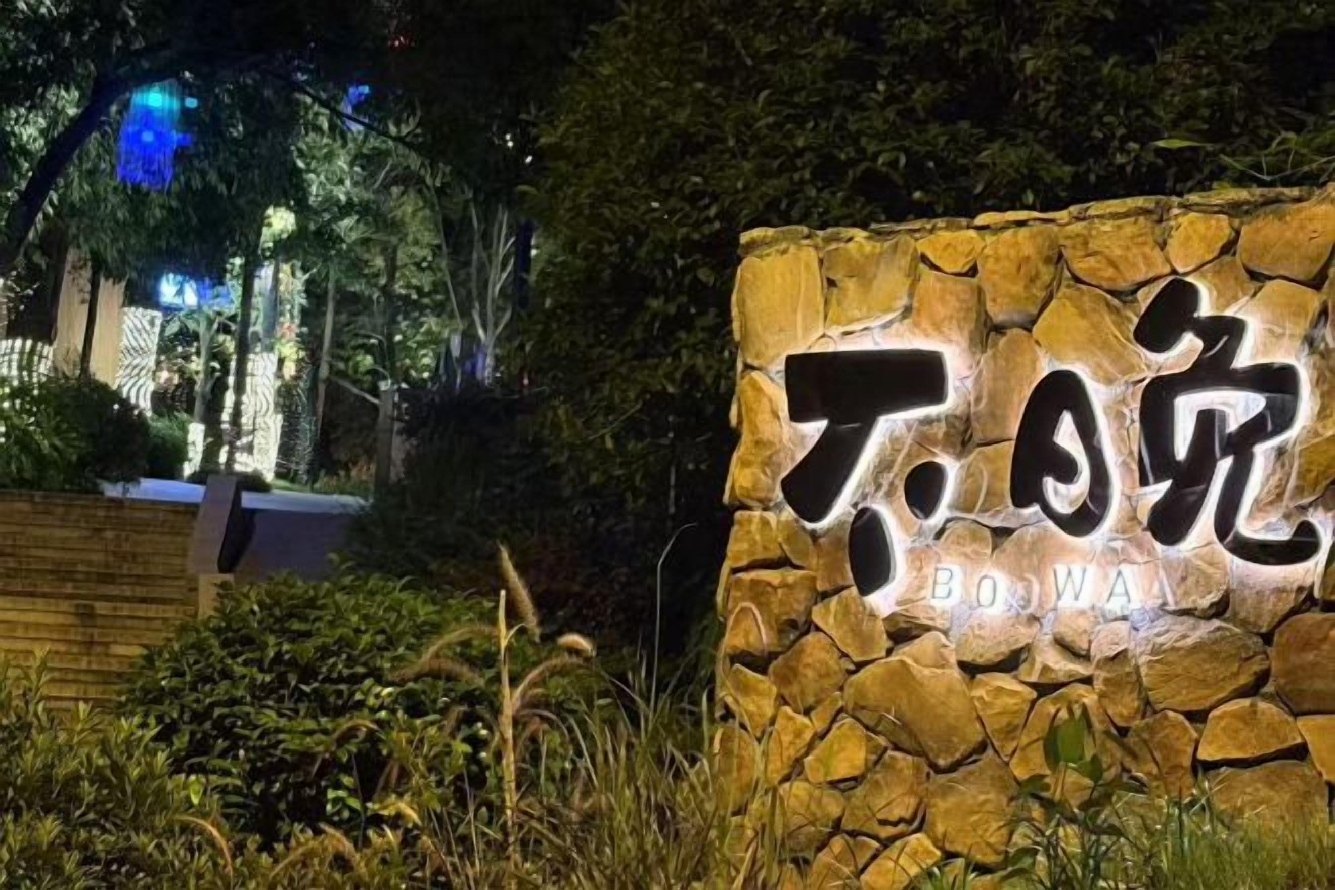 Un muro de piedra con texto iluminado, presumiblemente un letrero para un hotel o propiedad, rodeado de follaje.