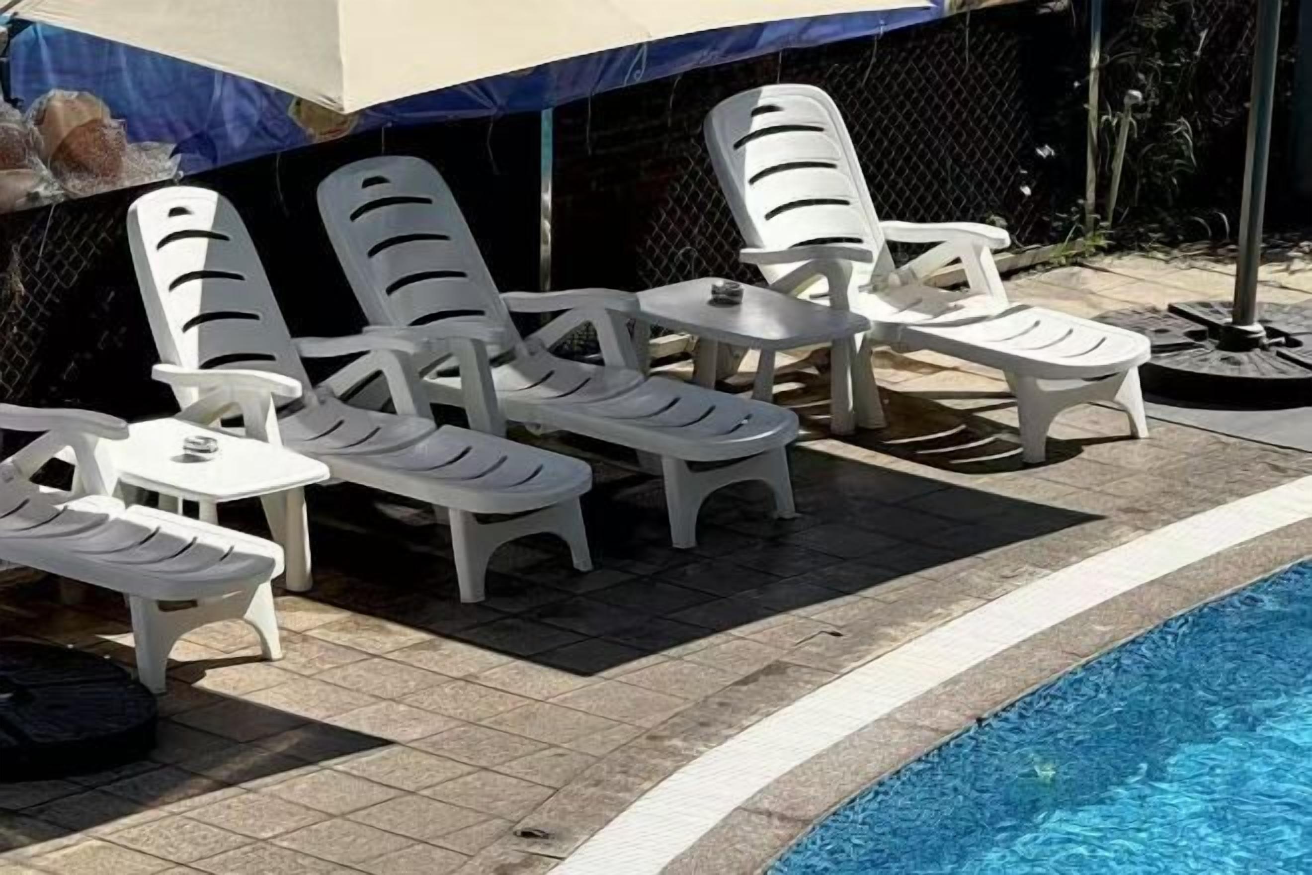 A poolside area with white plastic lounge chairs and a small table, surrounded by a tiled deck.