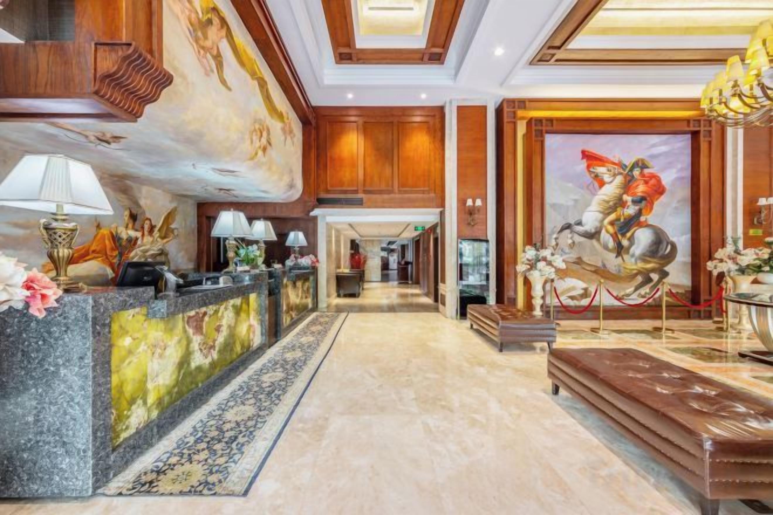 Um hall de hotel com uma mesa de recepção, uma escadaria imponente e um mural grande.