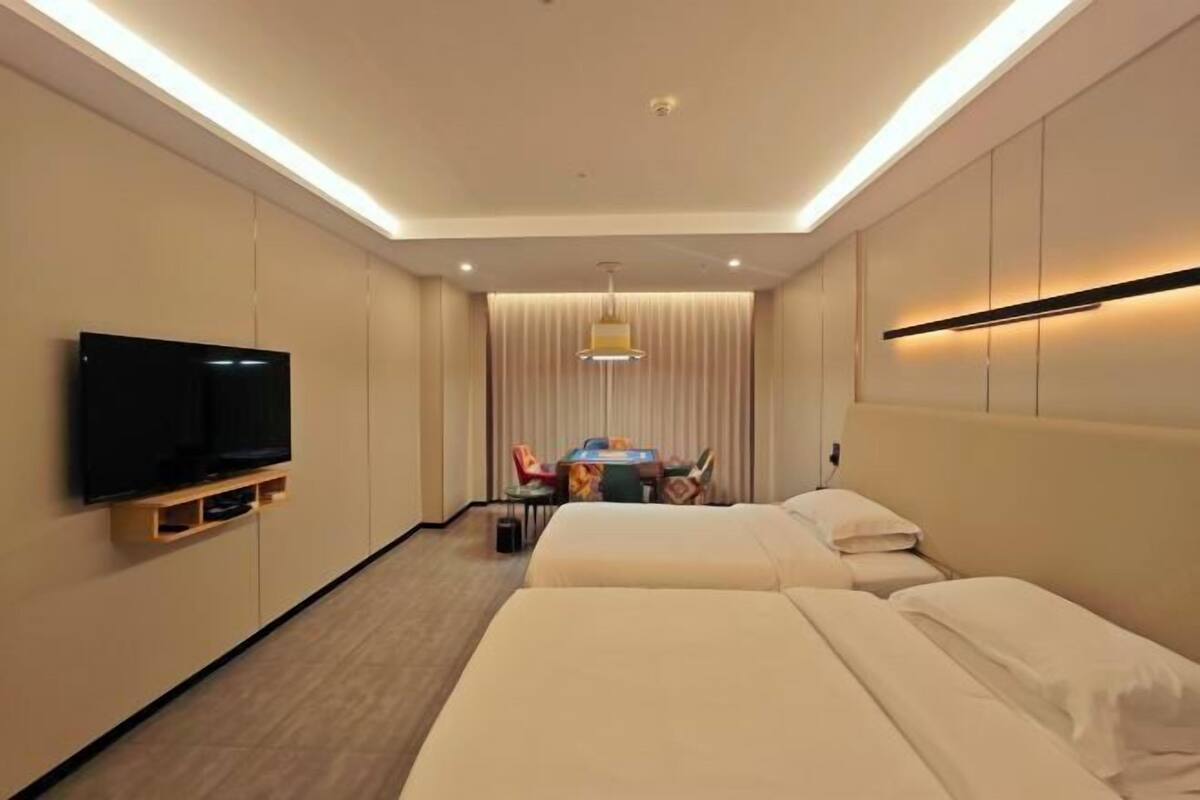 Habitación de hotel con dos camas, un televisor de pantalla plana montado en la pared, una mesa de comedor con sillas y una lámpara en el techo.
