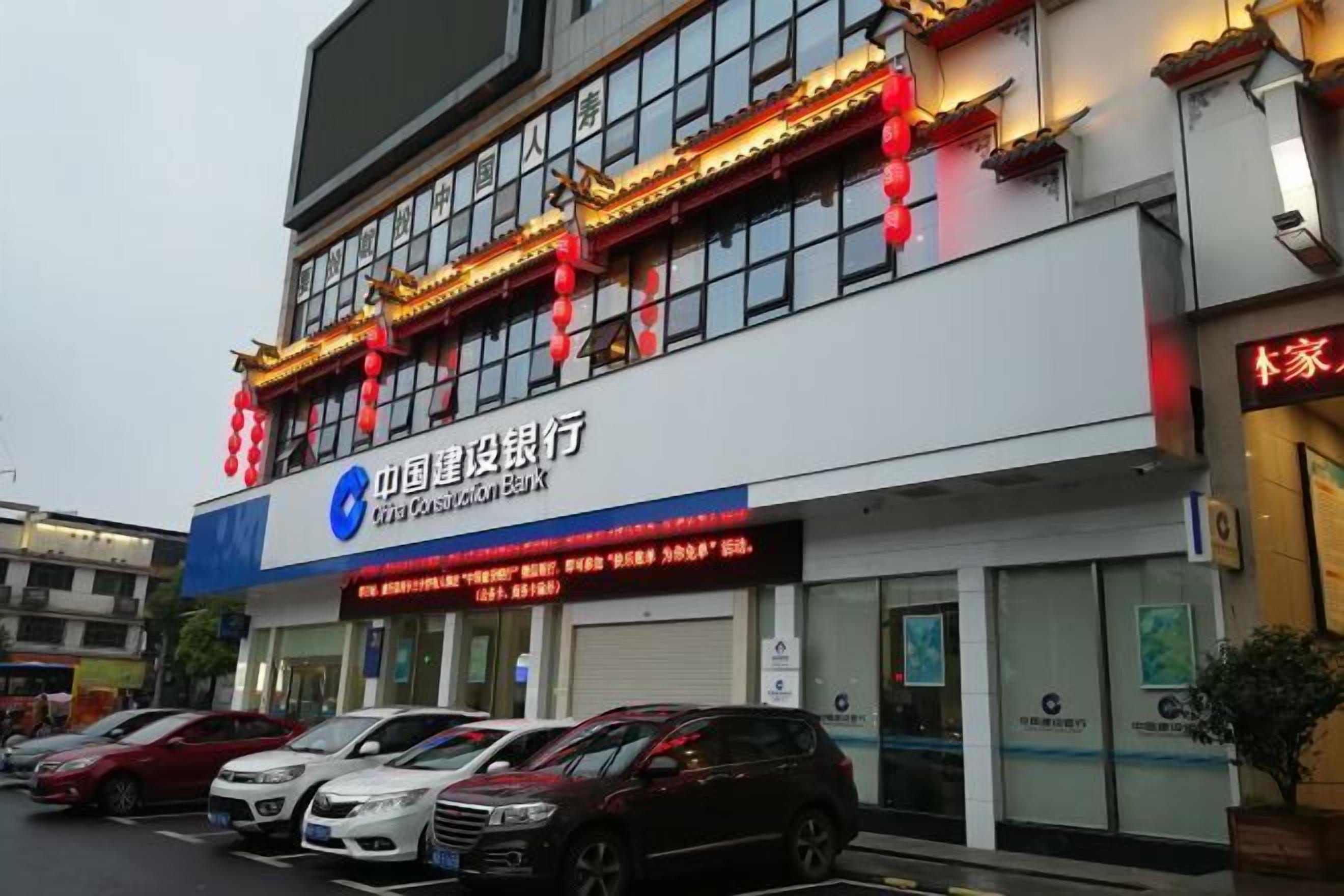 Ein Gebäude der China Construction Bank mit einer Werbetafel, abgestellten Autos und einer roten Laternenverzierung.