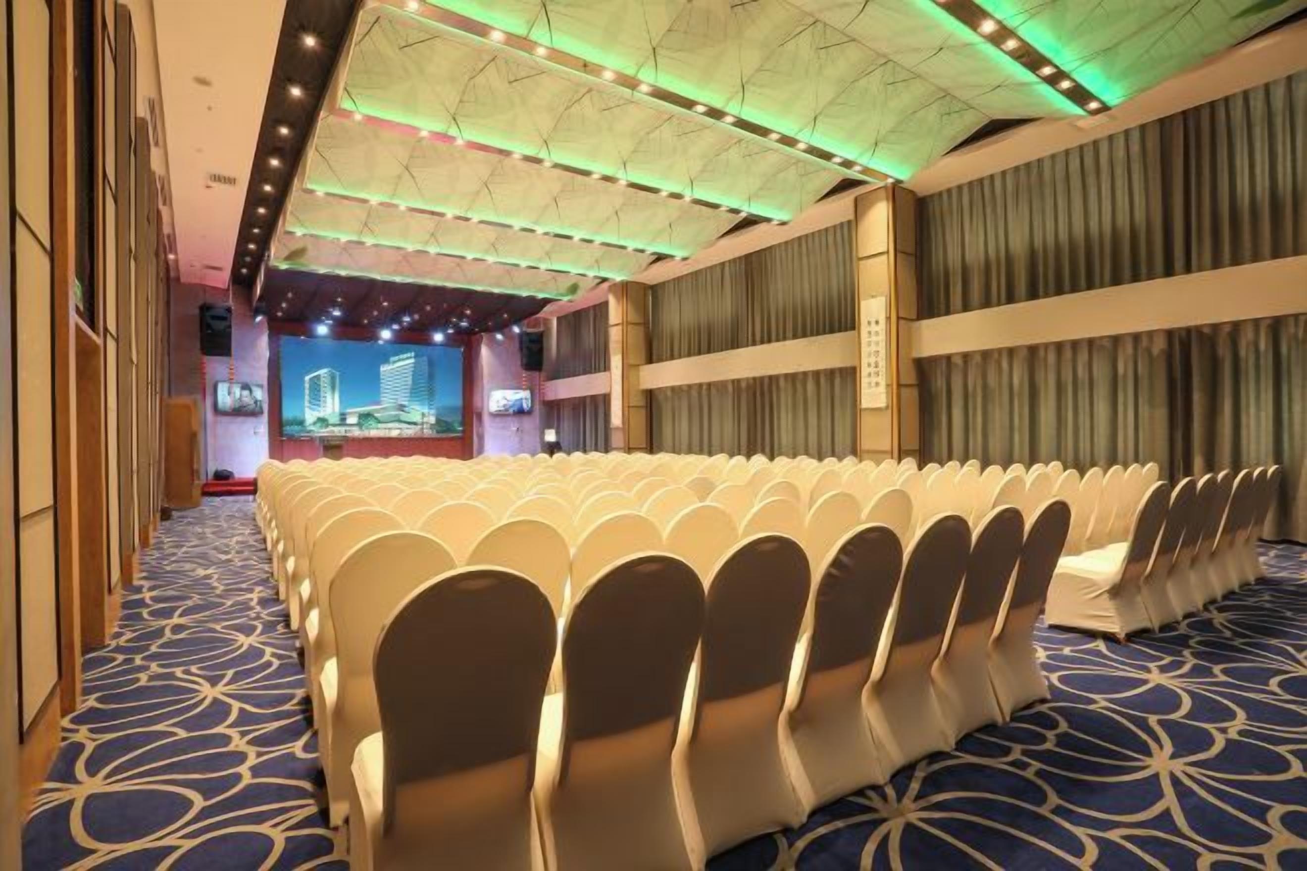 Un salón de conferencias con filas de sillas, una pantalla grande y una alfombra estampada.