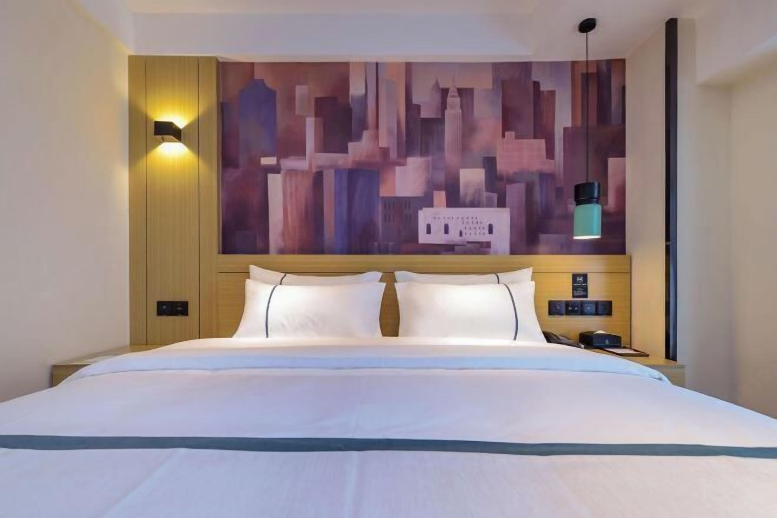 A modern hotel room with a large bed, a cityscape mural on the wall, and a nightstand with a lamp.