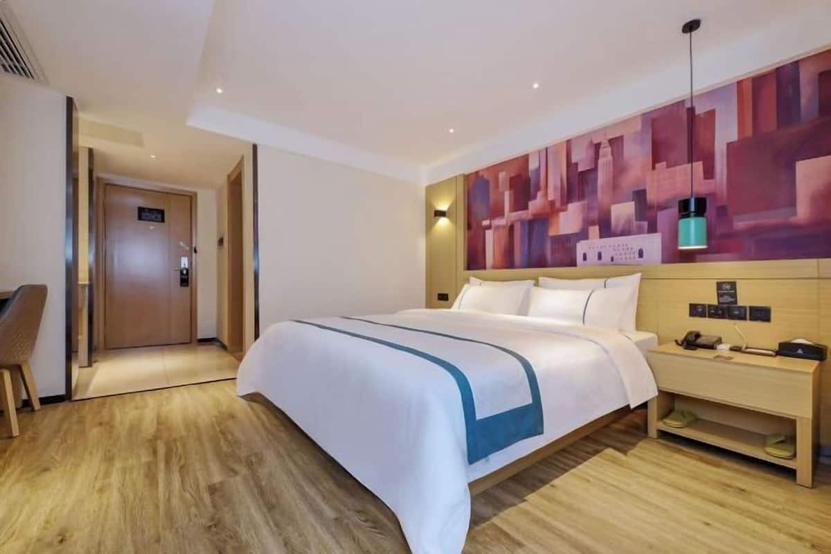 A hotel room with a large bed, a wooden nightstand, a desk, and a wall with a geometric patterned mural.