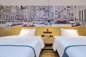 Habitación de hotel con dos camas, un cabecero de madera, una mesita de noche con teléfono y un mural en la pared que muestra una calle de la ciudad.