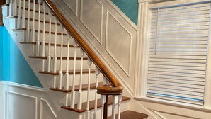 Uma escadaria com corrimãos brancos e degraus de madeira, conduzindo a um quarto com uma parede azul e uma janela com persianas.