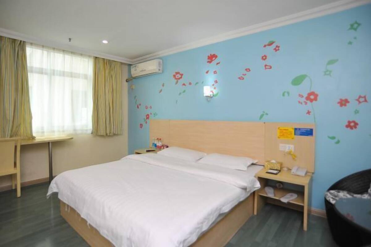 A hotel room with a bed, a bedside table, a chair, a window with curtains, and a wall with floral decals.