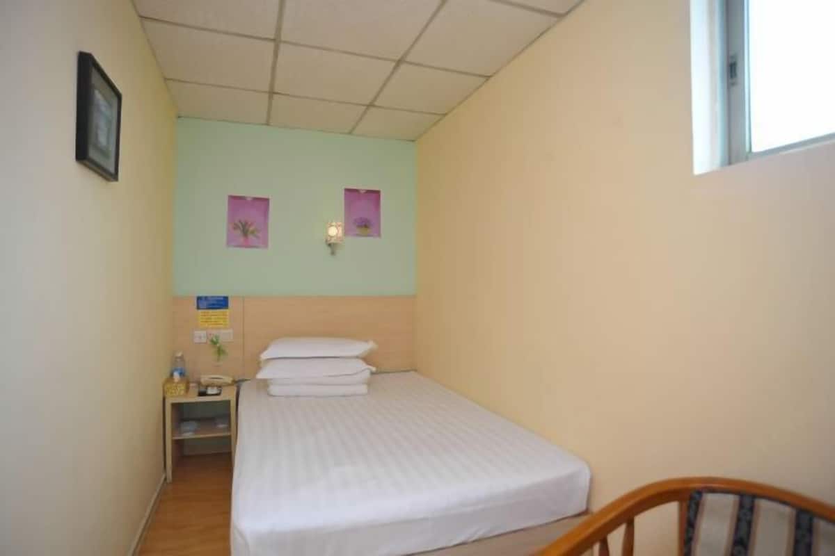 A small, single-bed room with a wooden headboard, a bedside table with a lamp, and a window.