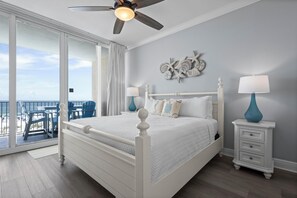 Relax in this serene hotel room featuring a large bed, ceiling fan, and sliding glass door leading to an outdoor area with a beach view.