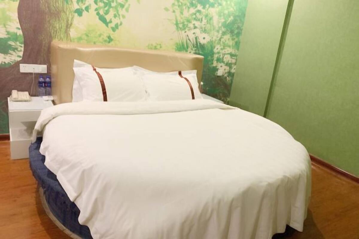 A neatly made bed with white bedding and brown accents, a nightstand with a phone and water bottle, and a wall mural of a tree and greenery.