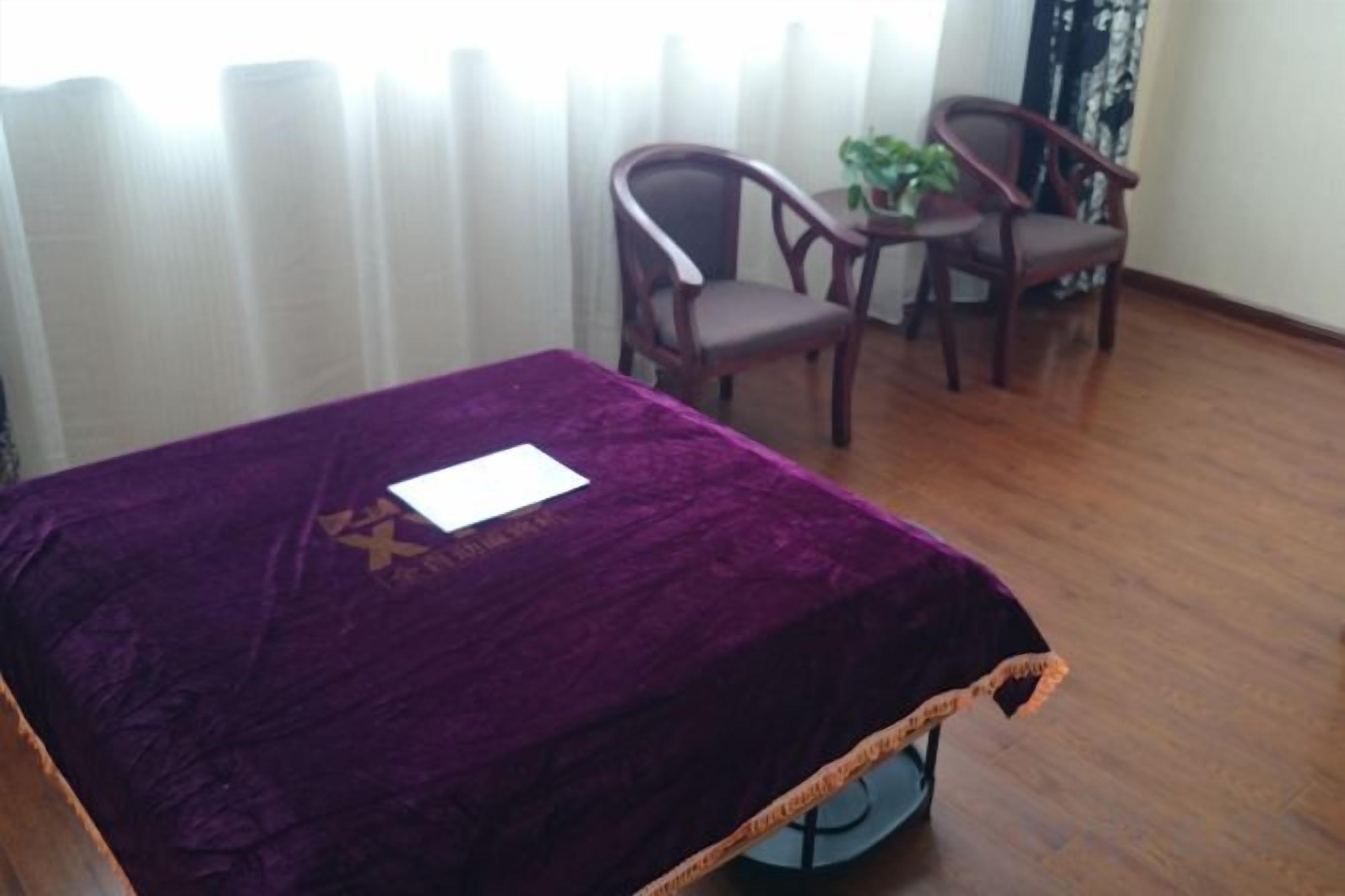 Ein Zimmer mit einem Bett, das mit einer violetten Decke bezogen ist, zwei Holzsesseln, einem kleinen Tisch mit einer Pflanze und einem Fenster mit Vorhängen.